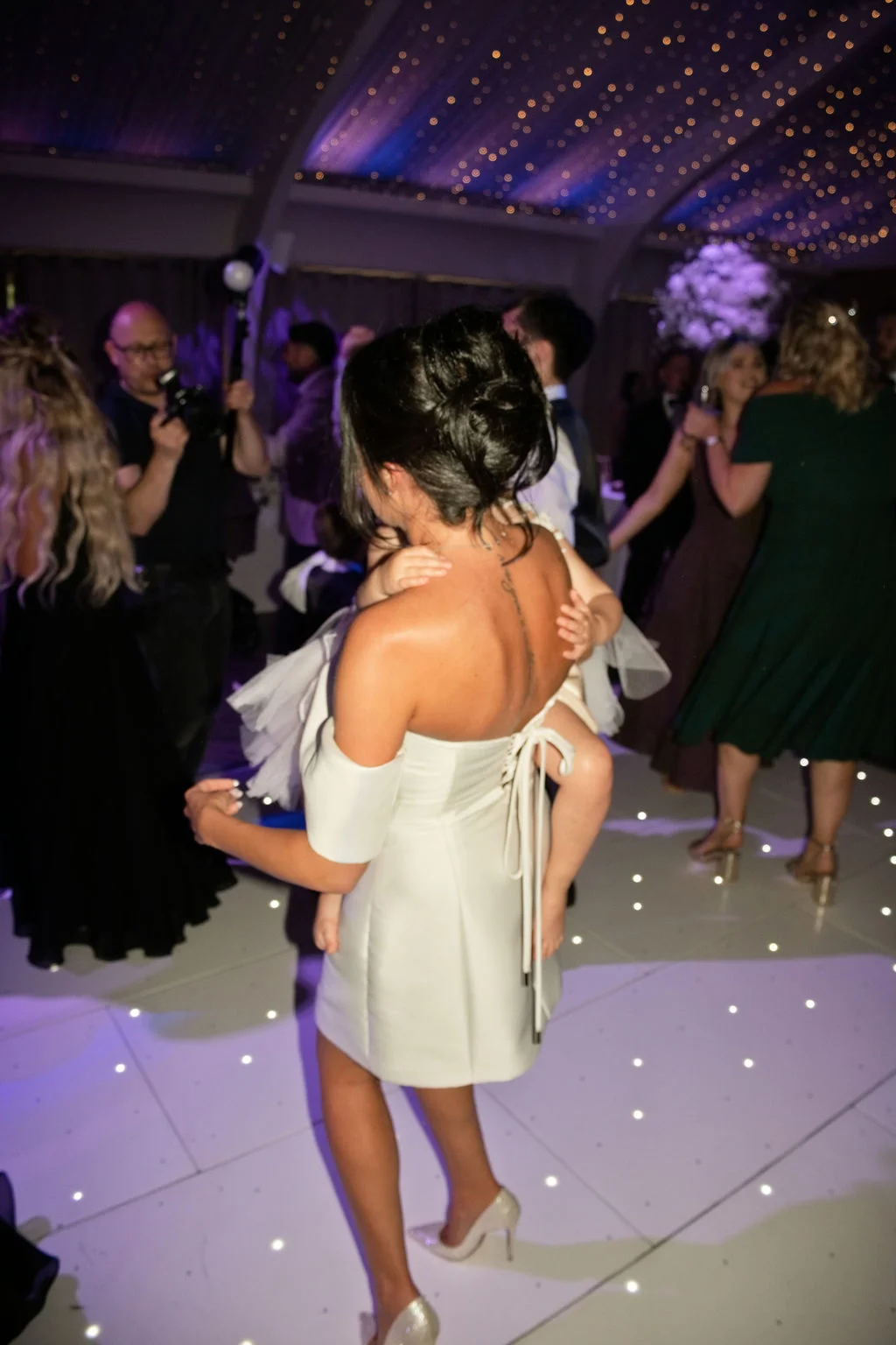 A guest in a short white off-the-shoulder reception dress carries a toddler on the dance floor beneath a sparkling fairy light canopy.