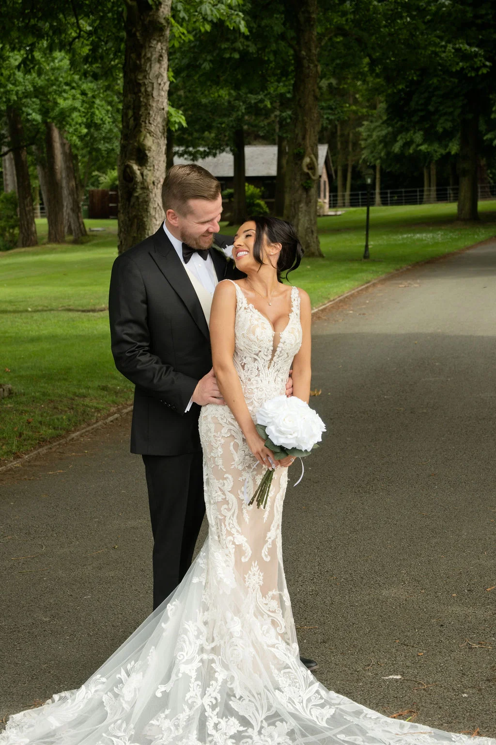 A newlywed couple embraces on a tree-lined driveway, with the smiling bride holding a white rose bouquet while looking up at her groom.