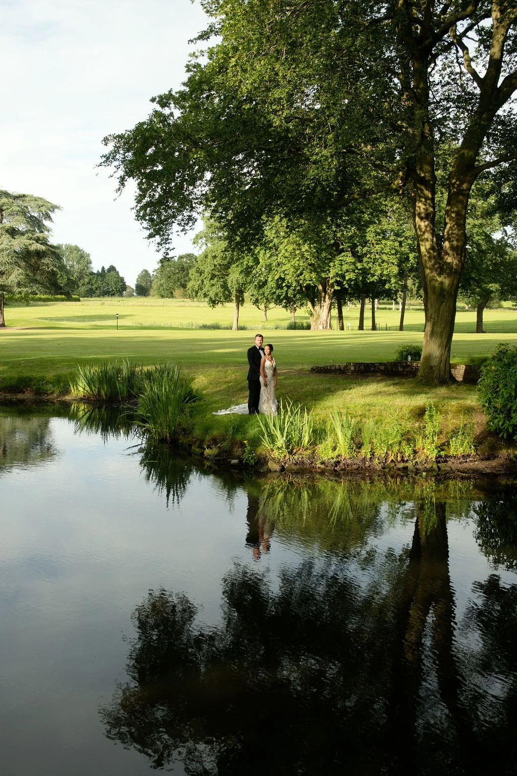 A newlywed couple stands by a tranquil lake under the shade of a large oak tree, surrounded by expansive manicured lawns.