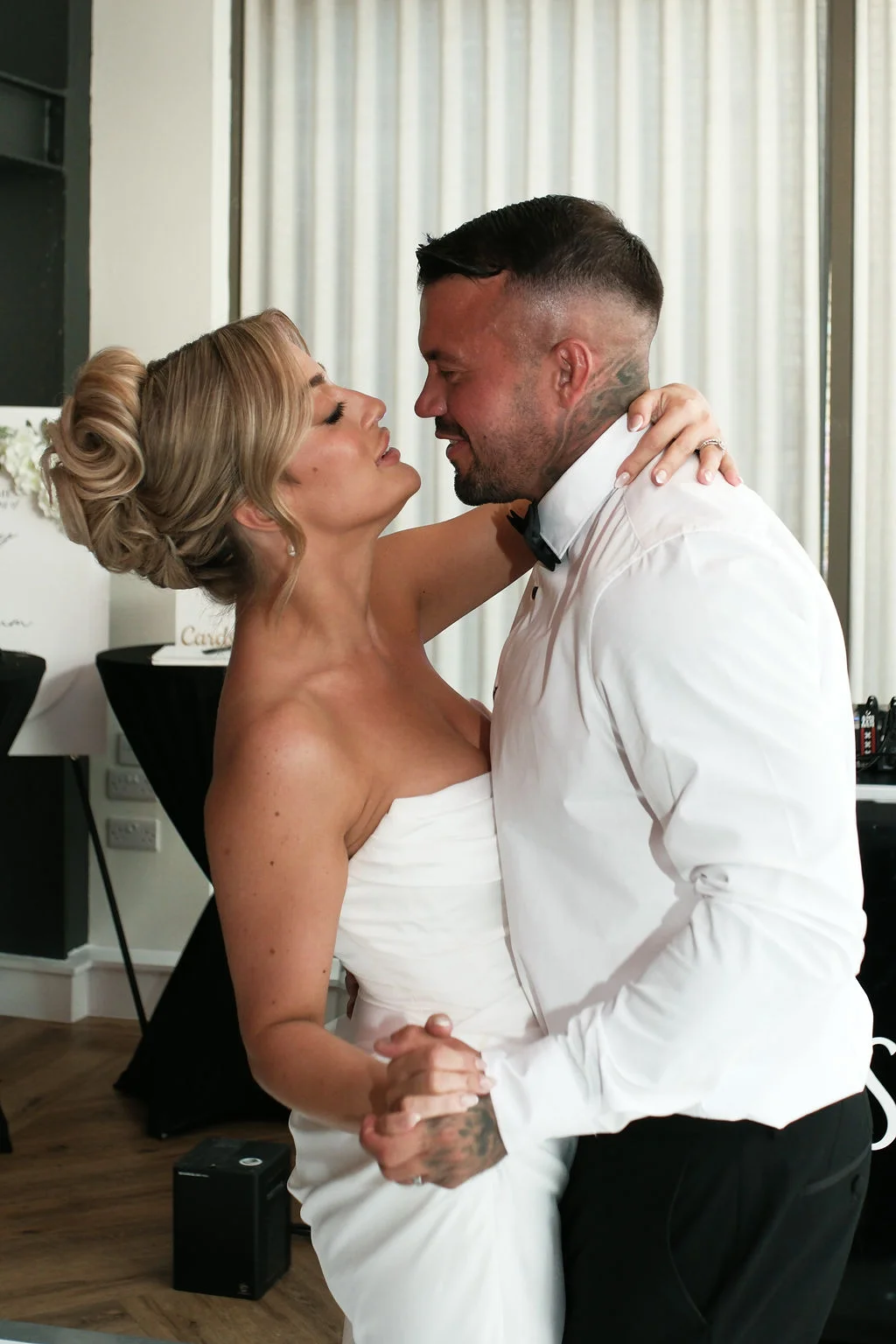 A newlywed couple share a tender first dance indoors, with the bride wearing an elegant strapless gown and a high bun while gazing up at her groom.