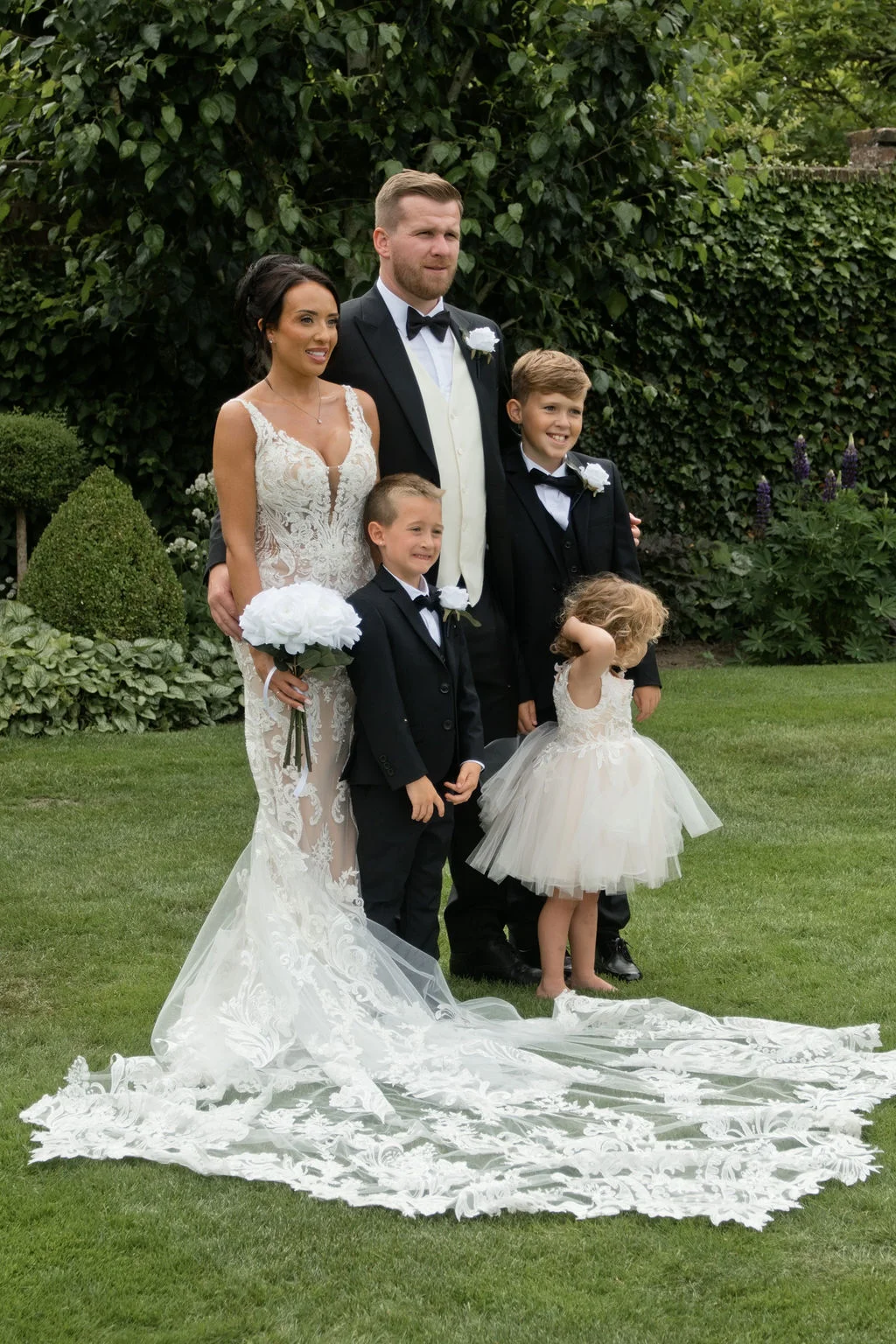 A newlywed family poses together on a manicured lawn with a lush green backdrop, featuring a bride in a lace mermaid gown with a dramatic train, a groom in a black tuxedo, and their children.