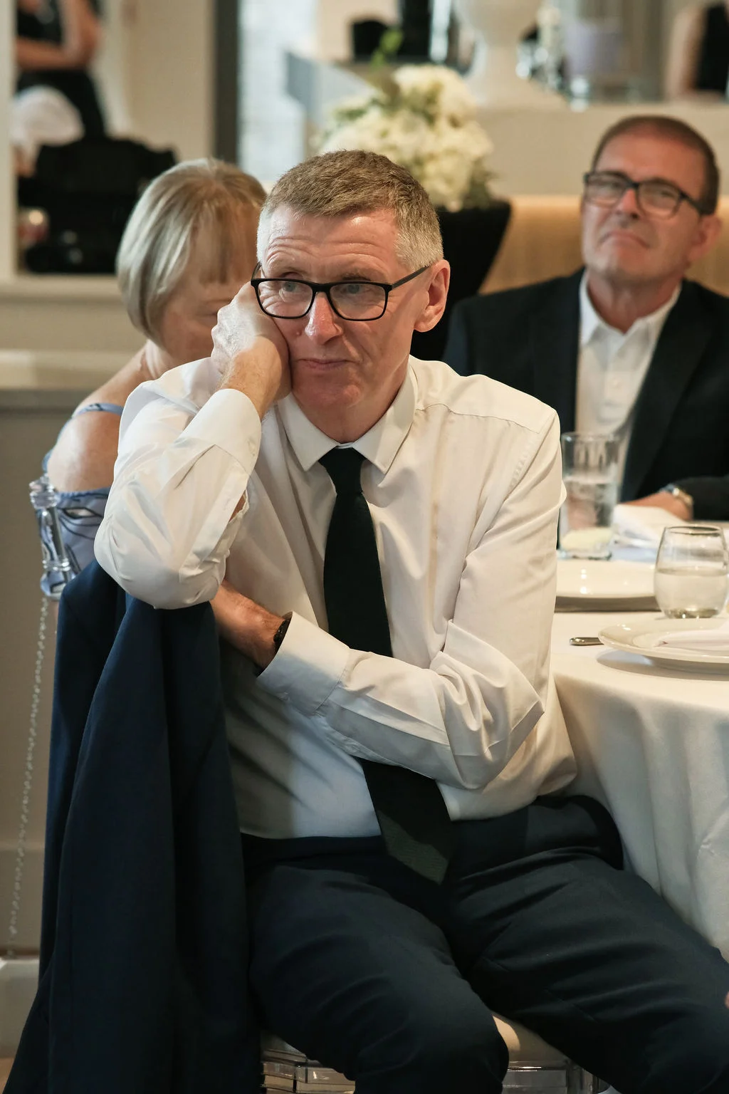 A guest sits attentively at a round reception table during wedding speeches, listening with his hand supporting his chin.