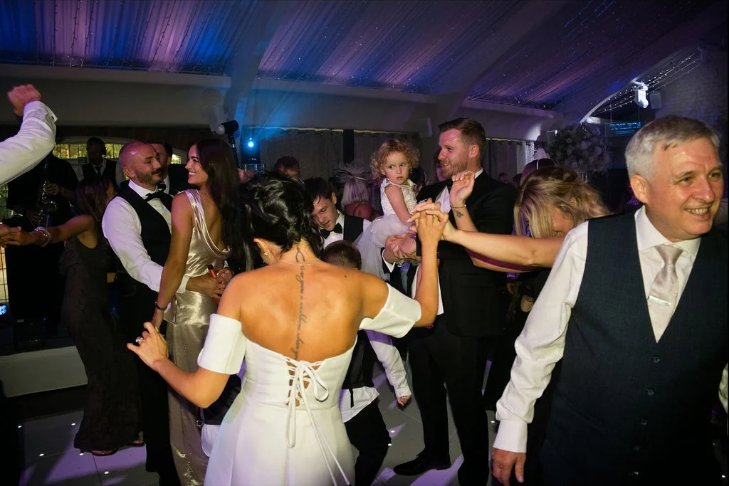 A lively evening reception captures guests of all ages dancing on a brightly lit dance floor underneath ceiling drapes and blue lighting.