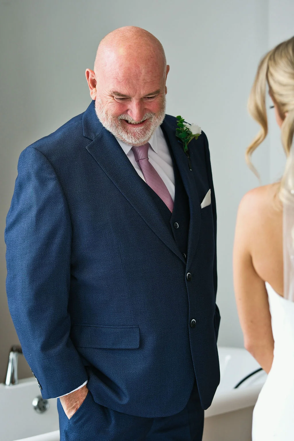 A proud and emotional father wearing a navy three-piece suit smiles lovingly as he shares a special first look moment with his bride daughter.