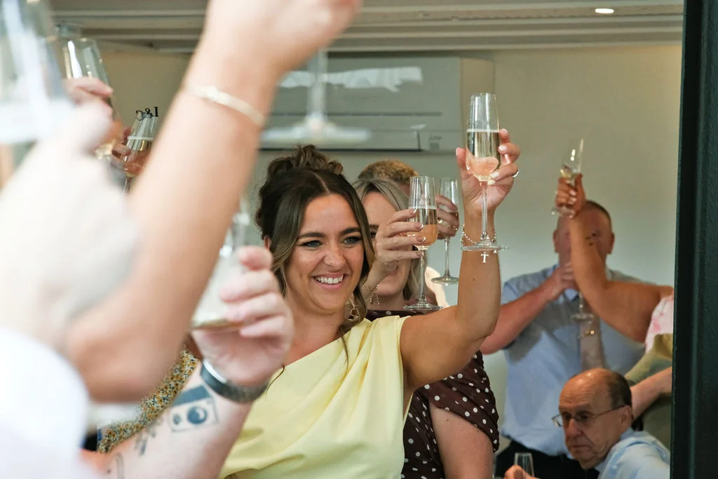 Wedding guests raise their champagne flutes in a cheerful toast while smiling and laughing during the celebration.