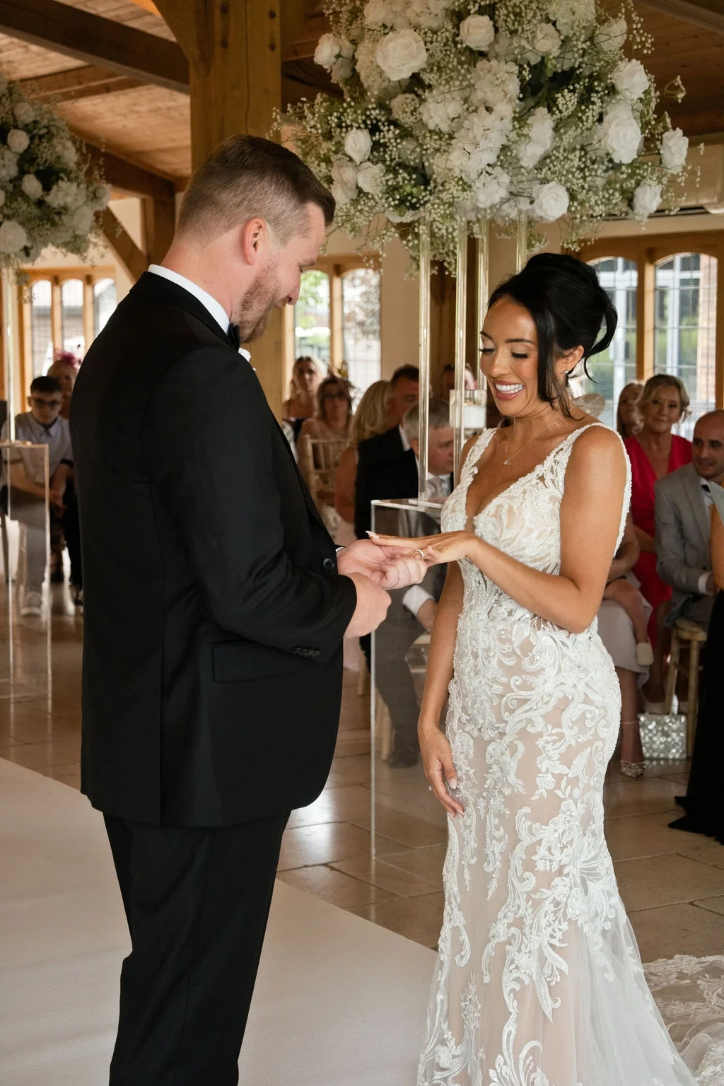 The groom places a wedding ring on the smiling bride's finger during their indoor ceremony beneath large white floral arrangements inside a rustic barn.