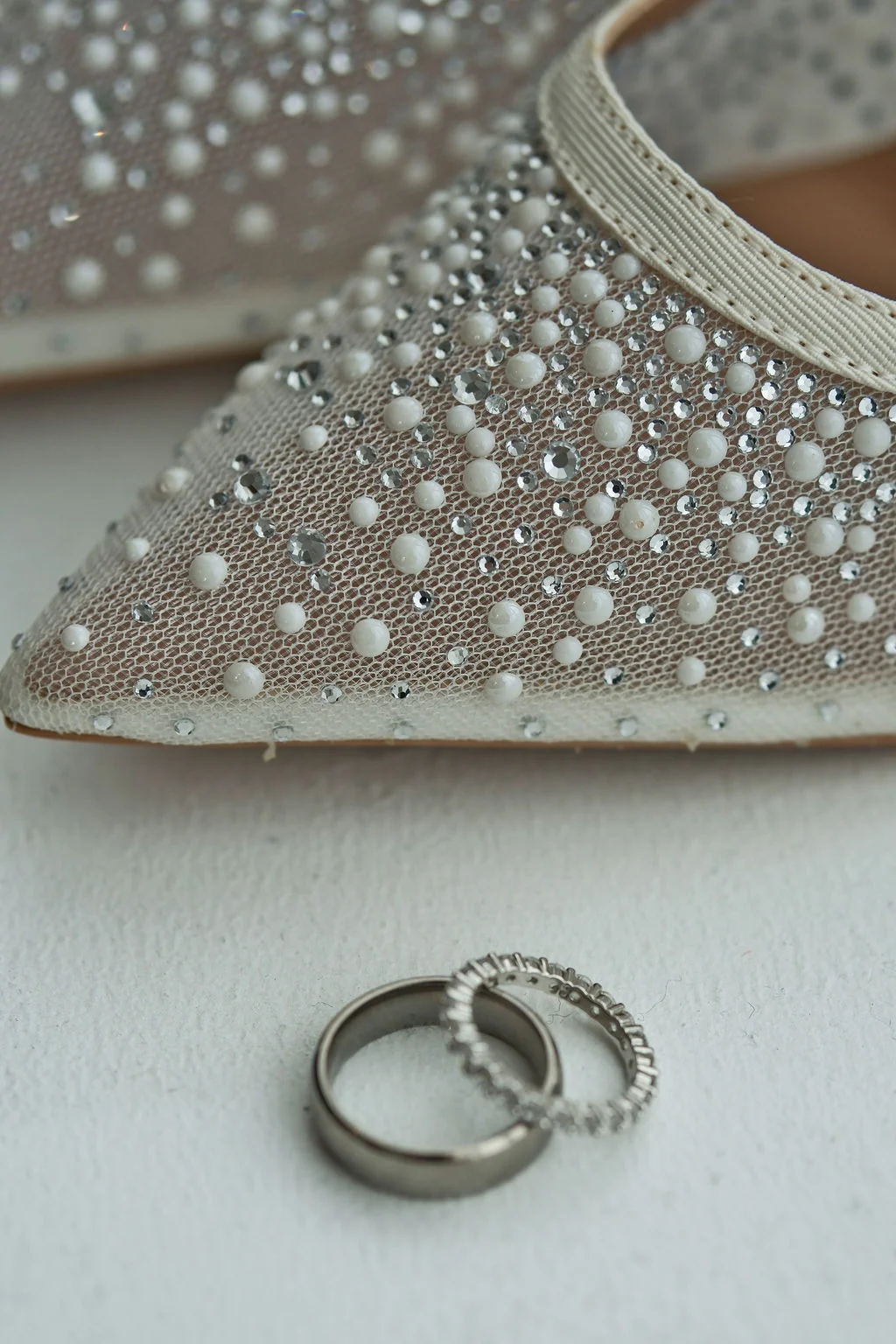A close-up shot of a bridal shoe heavily embellished with pearls and crystals resting just behind a pair of matching wedding bands.