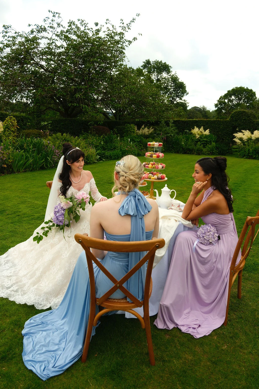 A bride in an off-the-shoulder lace gown and two bridesmaids in pastel blue and pink dresses sit around a table set for afternoon tea on a lush green lawn.