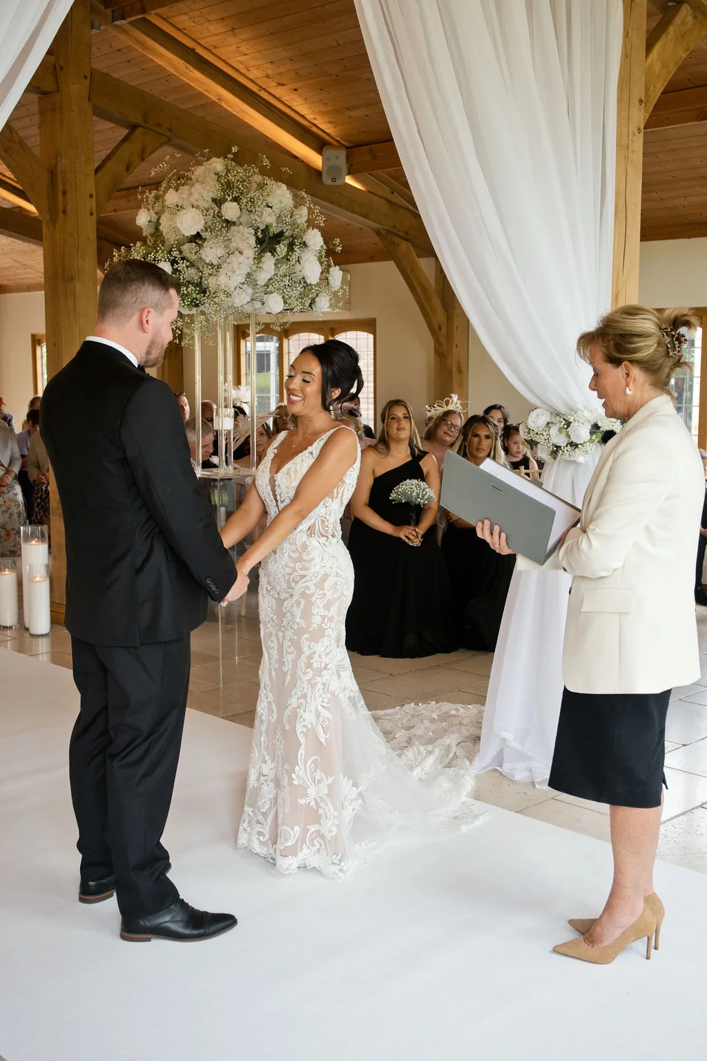 A bride and groom hold hands during a civil ceremony inside a light-filled oak framed barn, standing before a celebrant while guests watch from seating behind them.