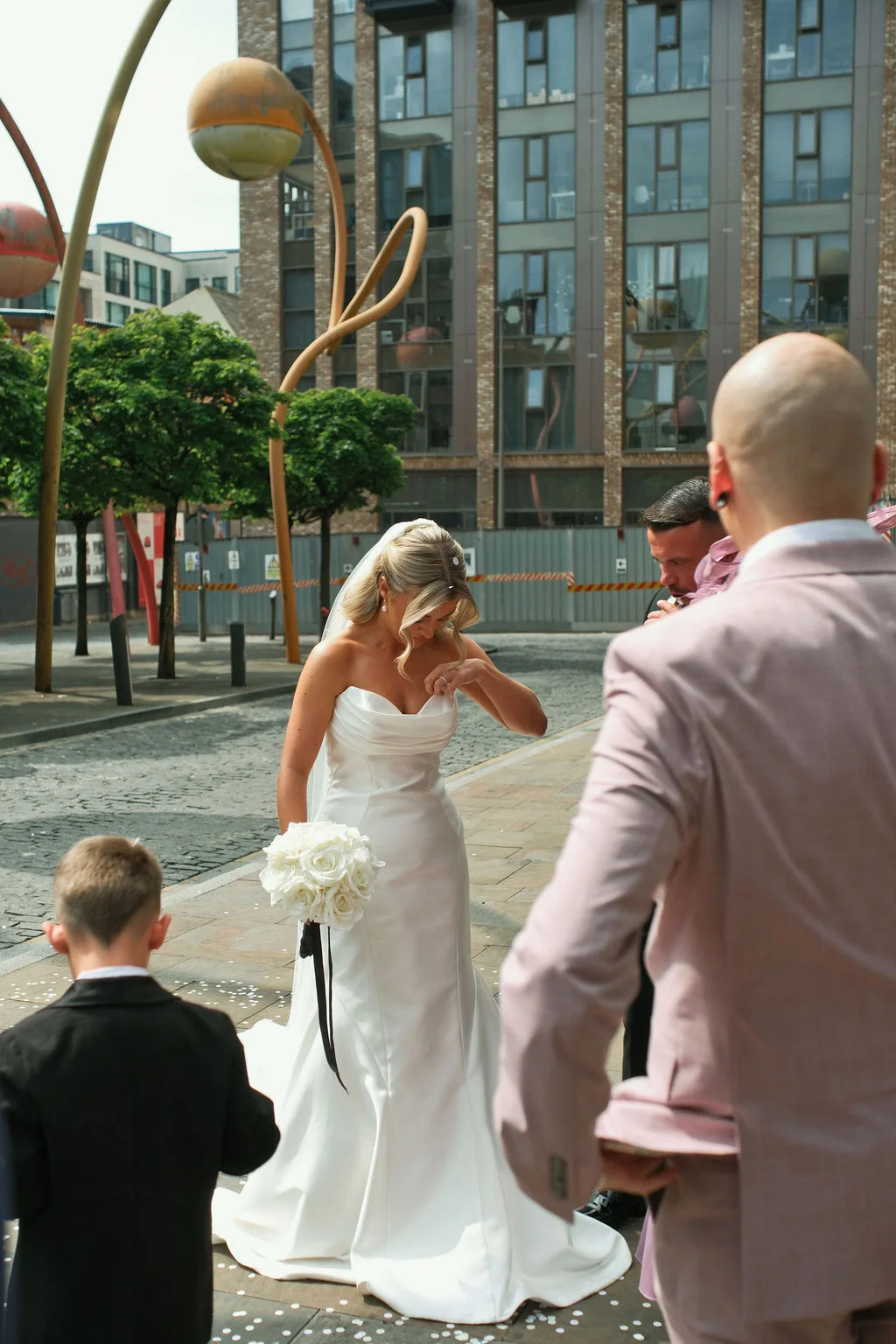 A stunning bride in a strapless satin gown holding a white rose bouquet stands on an urban cobbled street while guests look on.