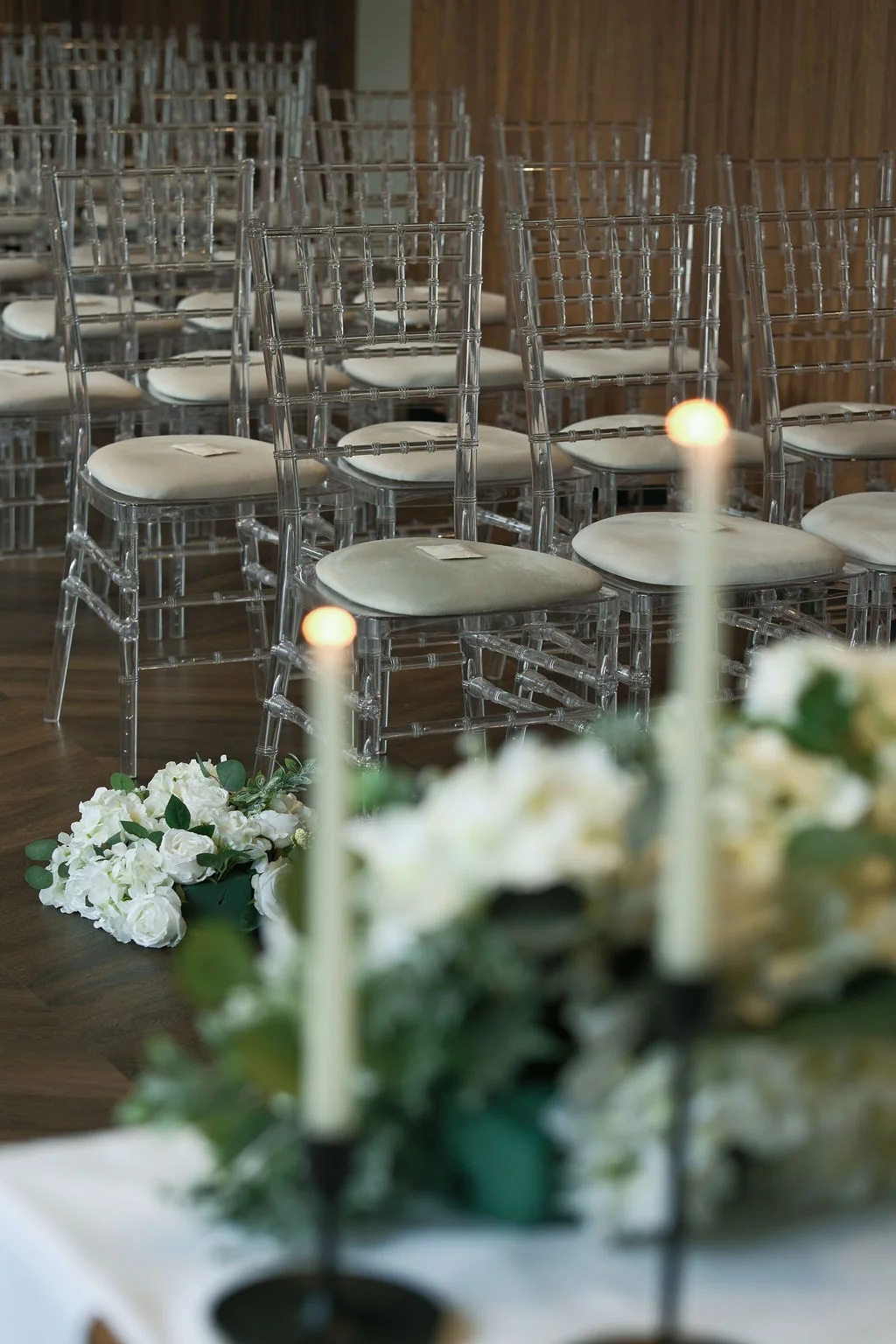 An empty wedding ceremony space features clear ghost chairs arranged neatly in rows, with a soft-focus floral arrangement and glowing taper candles in the foreground.