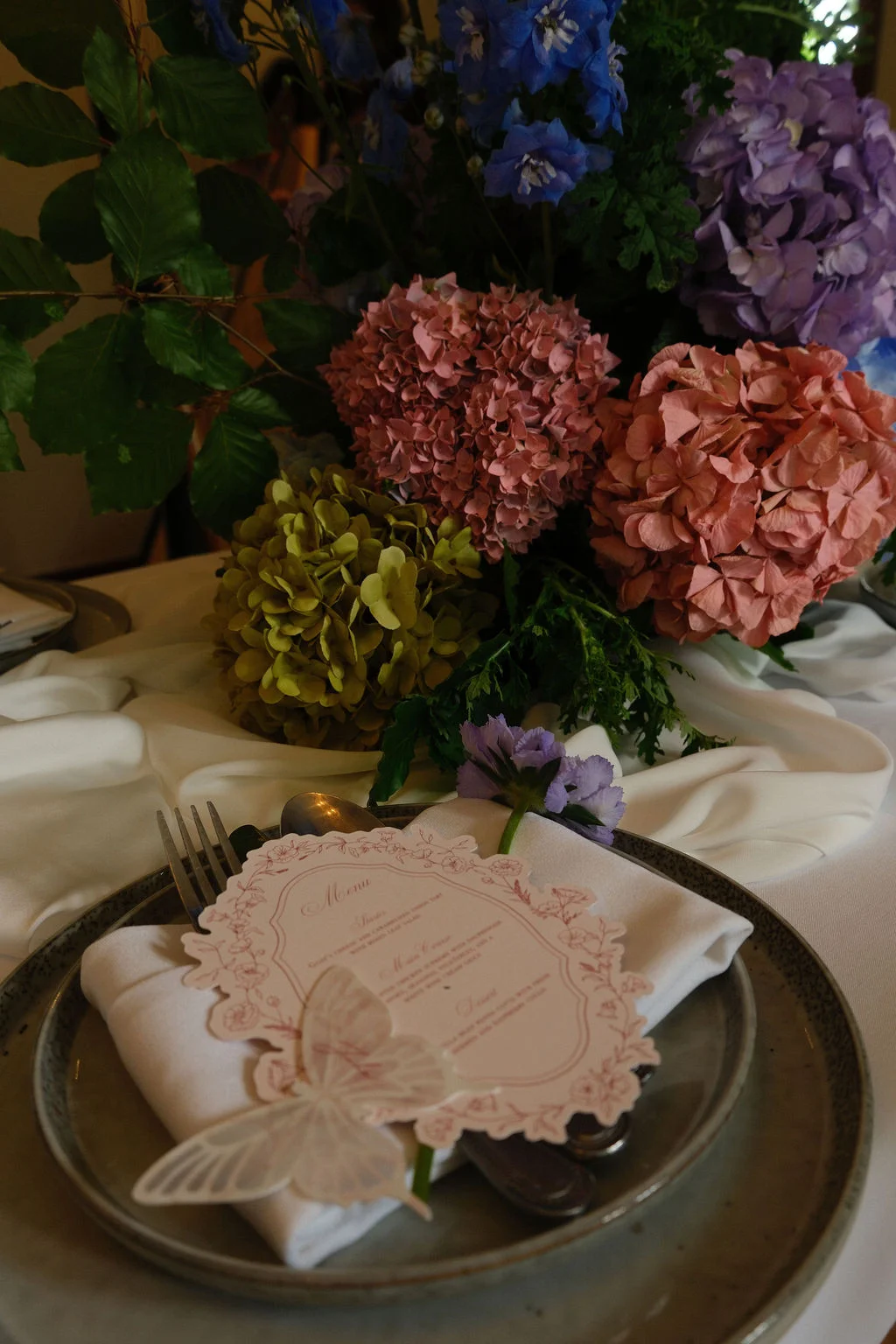 A close-up of a wedding place setting featuring a decorative menu card and butterfly detail resting on a white napkin over layered plates, set against a backdrop of lush hydrangeas and delphiniums.