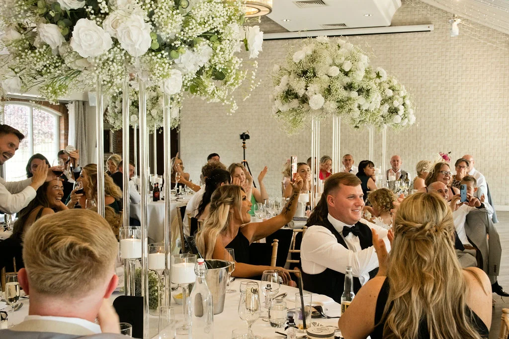 A groom delivers a speech at the top table during a wedding reception, surrounded by guests laughing and applauding under tall white floral centrepieces.