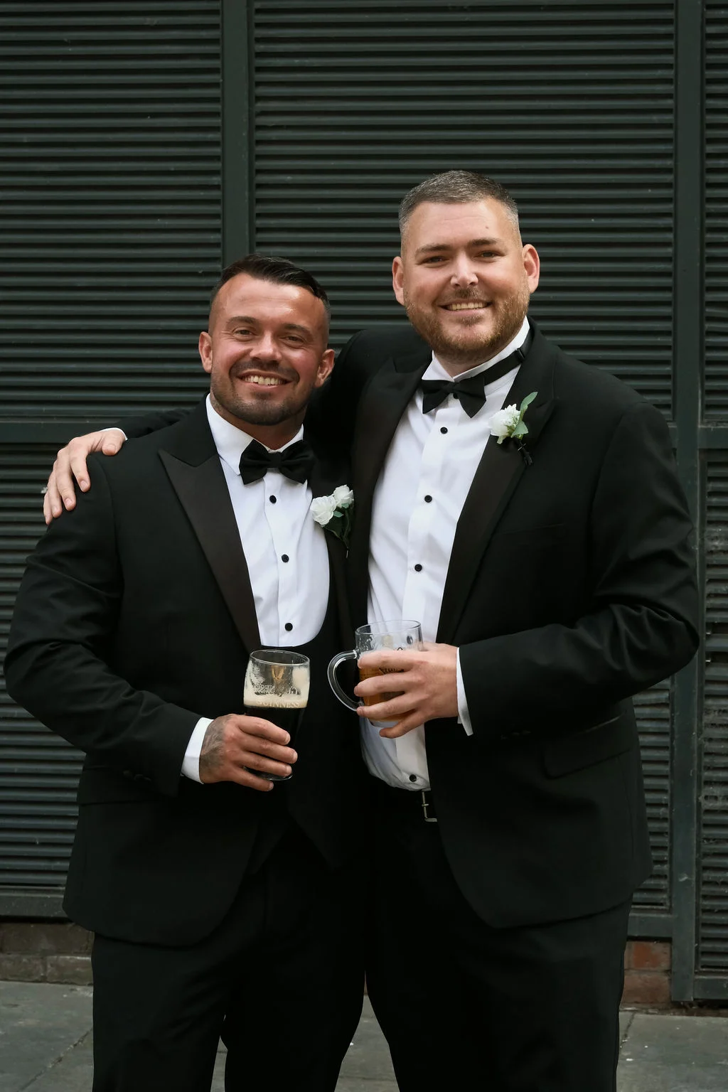 Two smiling grooms in classic black tuxedos pose together during the drinks reception, each holding a refreshing pint of beer.