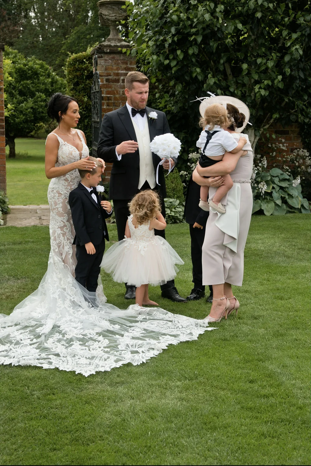 A newlywed couple and their family stand on a manicured lawn outside an estate, with the bride wearing a stunning lace gown featuring an elaborate train.