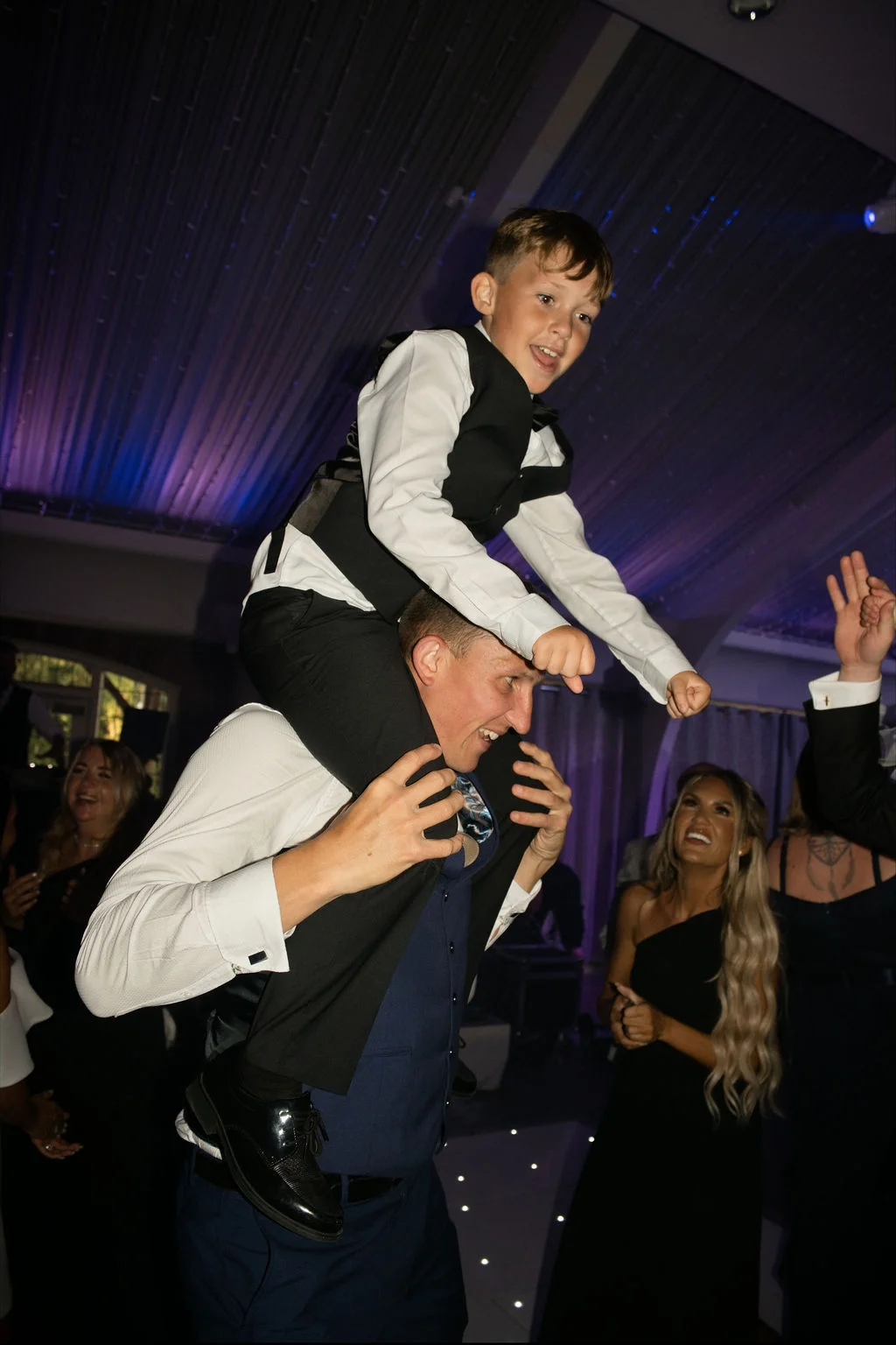 A young page boy is carried high on someone's shoulders on a lively evening dance floor illuminated by disco lighting.