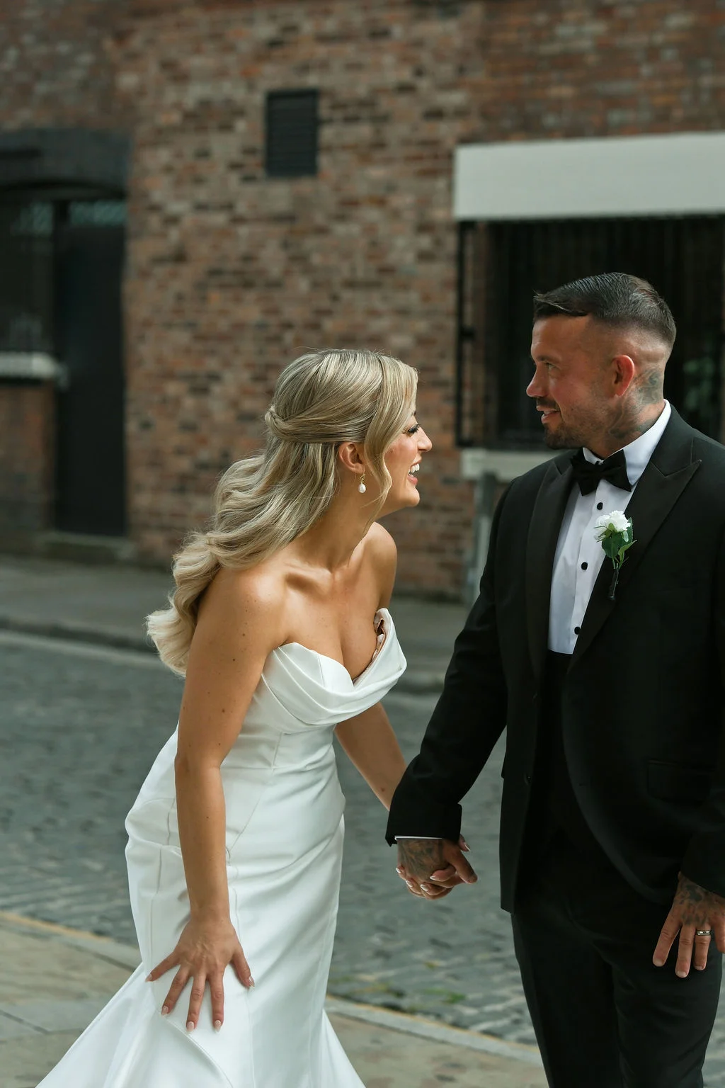 A newlywed couple shares a joyful laugh while walking hand in hand down a cobbled city street with an exposed brick backdrop.