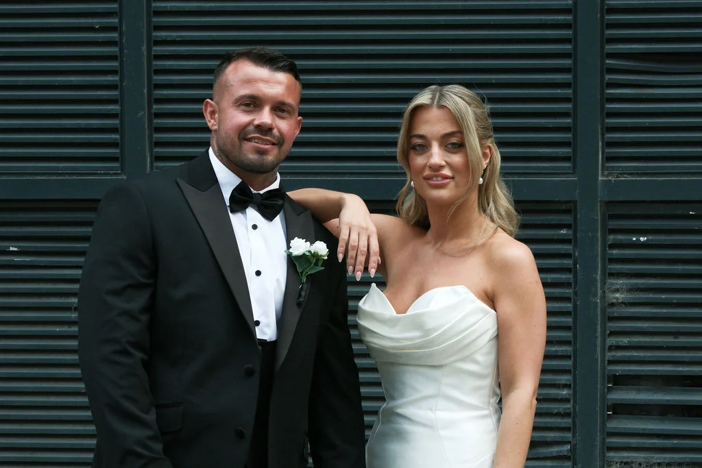A newlywed couple poses together in front of a dark slatted background, with the groom in a classic black tuxedo and the bride in a strapless satin gown.