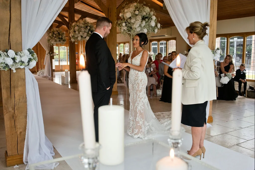 A bride and groom exchange rings during their wedding ceremony inside a bright oak-framed barn decorated with white floral arrangements and candles.