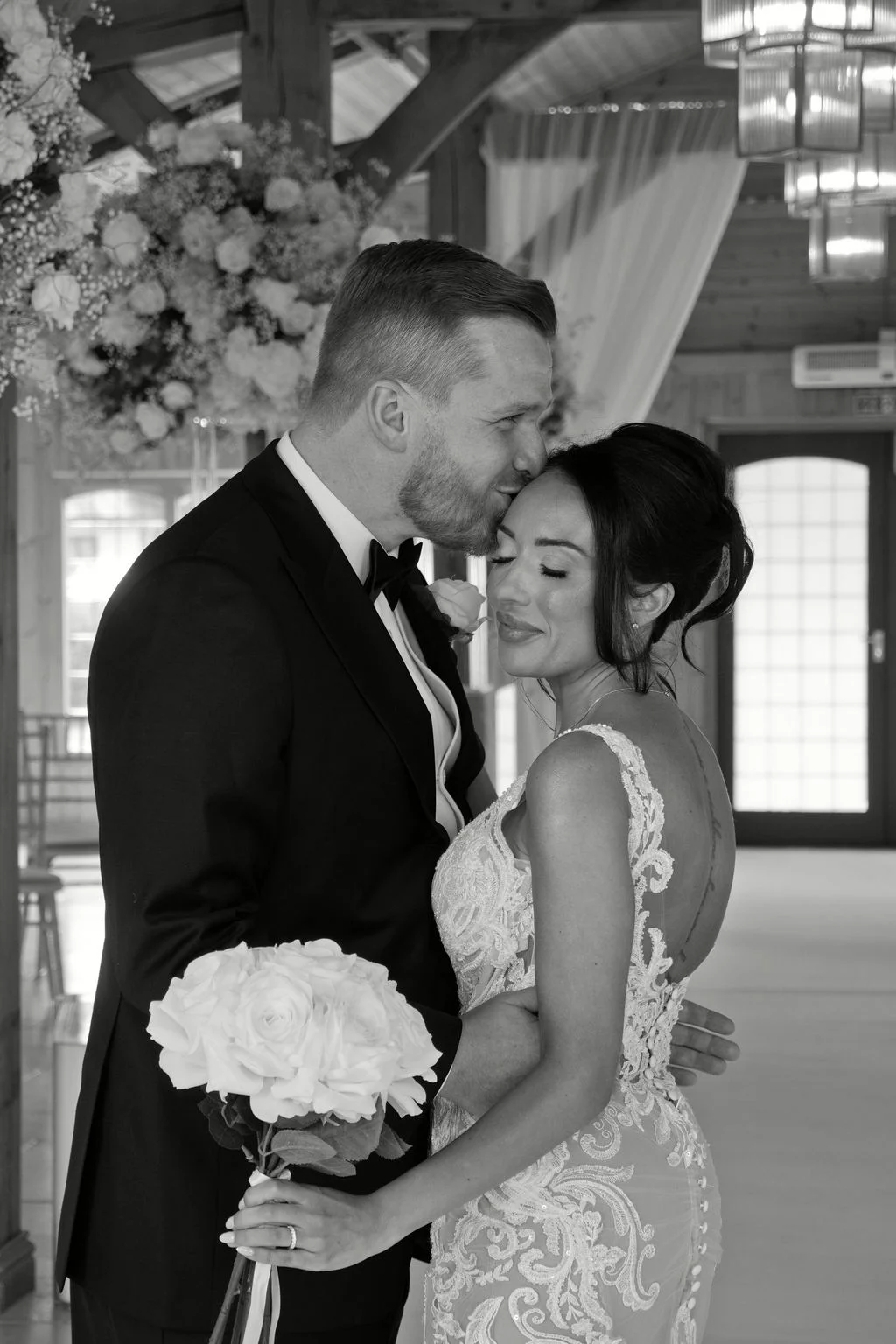 A groom gently kisses the bride on the forehead inside a rustic barn venue while she holds a white rose bouquet.