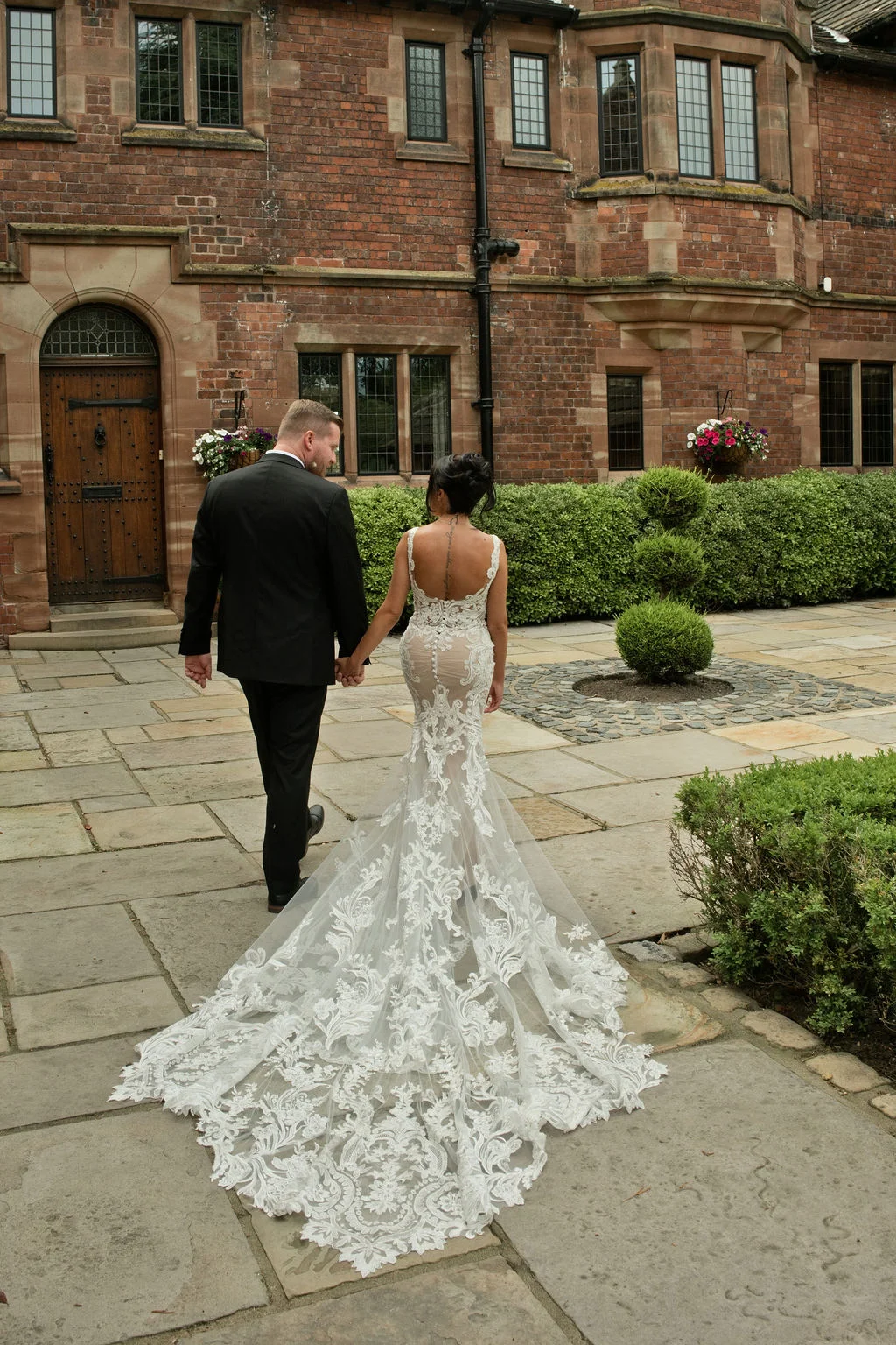 A newlywed couple holds hands and walks away across a stone courtyard outside a historic red brick manor house, showcasing the bride's dramatic lace gown with a long train.