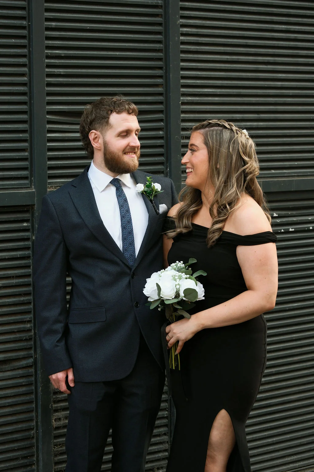 A smiling bride in a black off-the-shoulder dress holding a white rose bouquet, looking at her groom who wears a charcoal suit against an industrial slatted background.