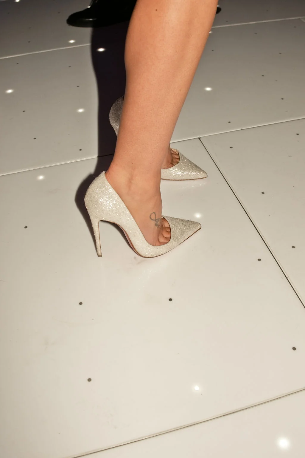 Close-up shot of a bride's foot wearing a sparkly high heel on a white lit dance floor, showing a small tattoo on the ankle.