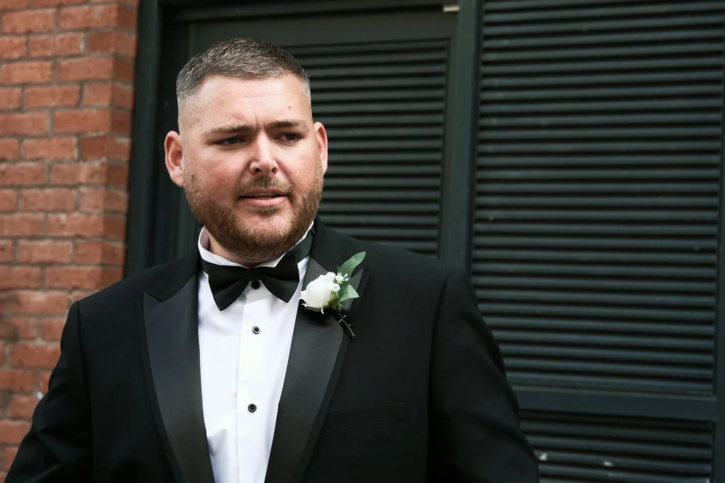 A close-up portrait of a groom wearing a classic black tuxedo with a white boutonniere against a brick wall backdrop.