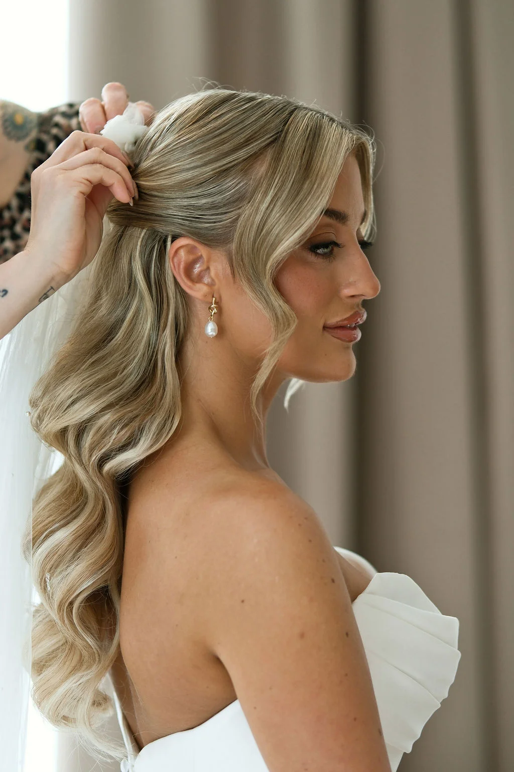A close-up profile shot of a blonde bride having her half-up hair styled with loose waves and delicate pearl earrings while wearing a strapless ruched wedding gown.