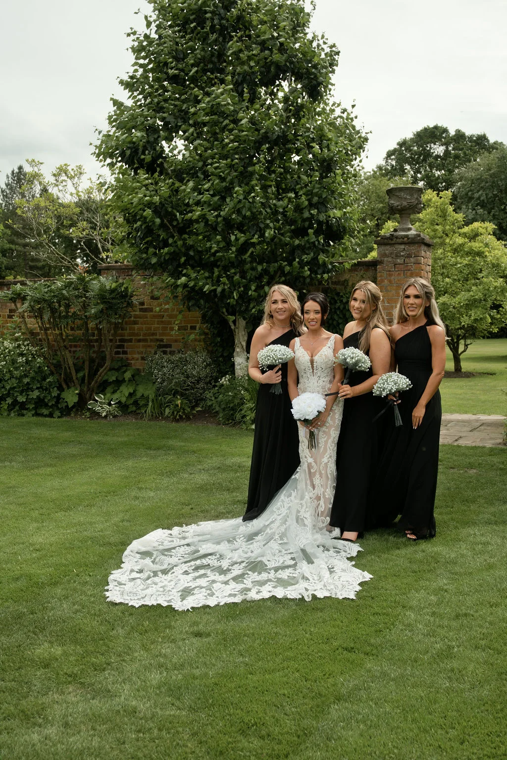 A stunning bride in an intricate lace gown with a dramatic cathedral-length train poses on a manicured lawn with four bridesmaids dressed in elegant black evening gowns, holding matching bouquets.