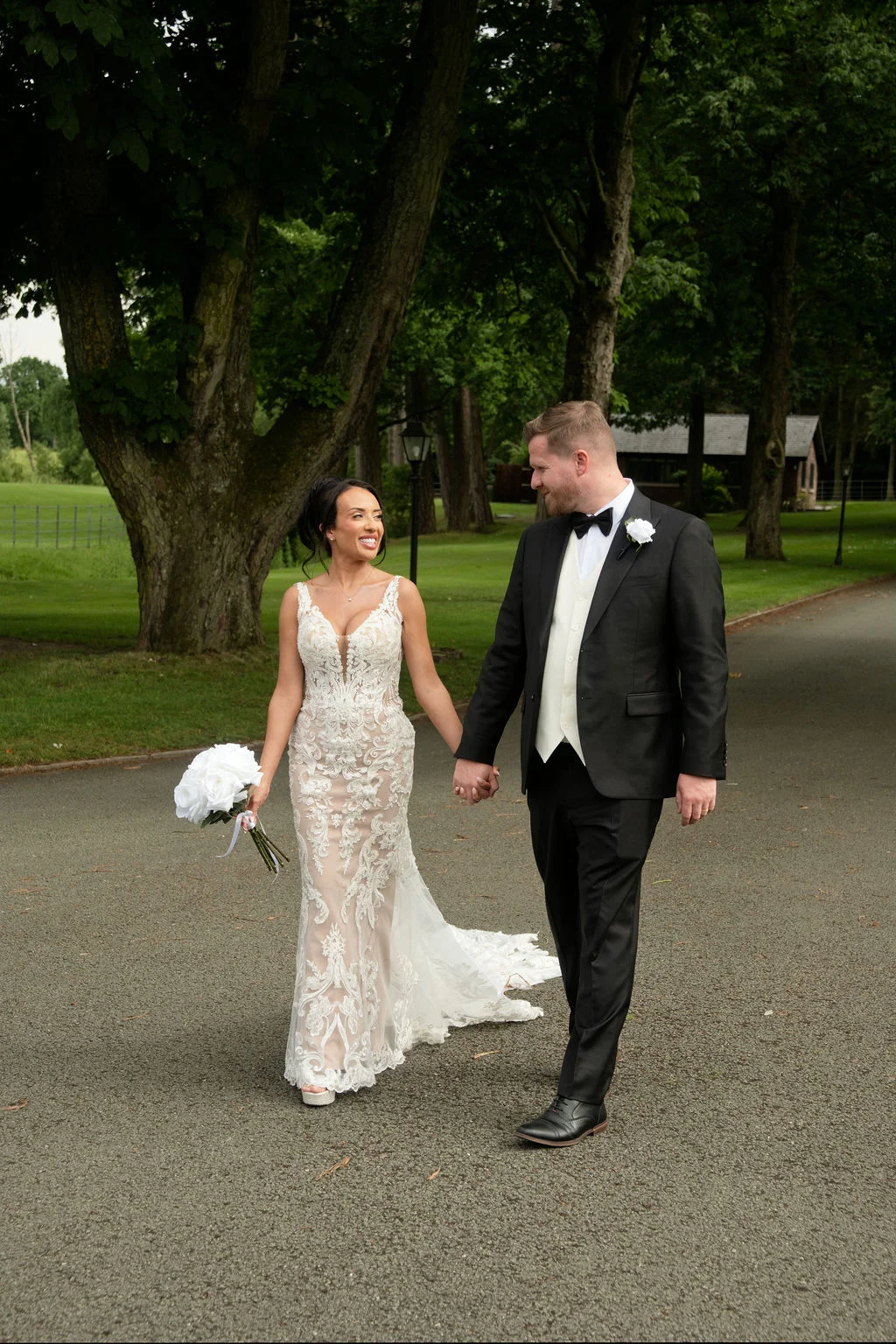 A smiling newlywed couple walks hand in hand along a paved driveway flanked by mature trees, with the bride wearing a fitted lace gown and holding a white bouquet while the groom wears a smart tuxedo.