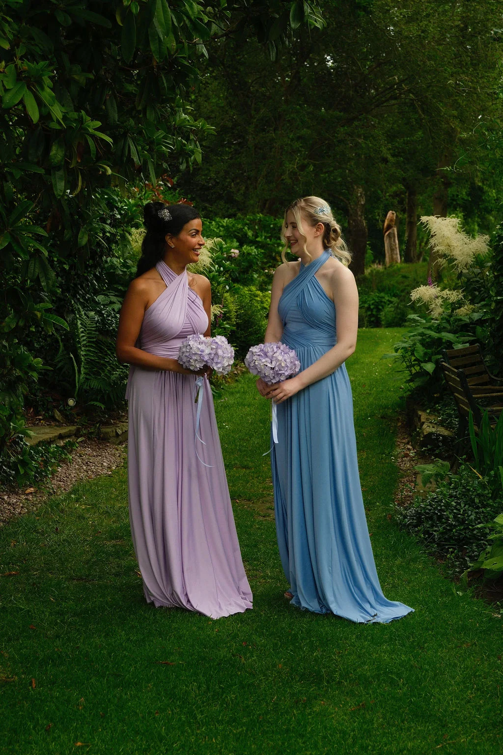 Two bridesmaids in pastel halterneck dresses stand on a lush green lawn holding matching hydrangea posy bouquets while chatting and smiling outdoors.