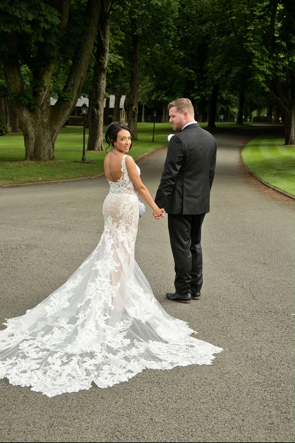 Newlyweds walking hand in hand down a paved path lined with tall trees, with the bride looking back over her shoulder wearing an intricate lace gown featuring a dramatic train.