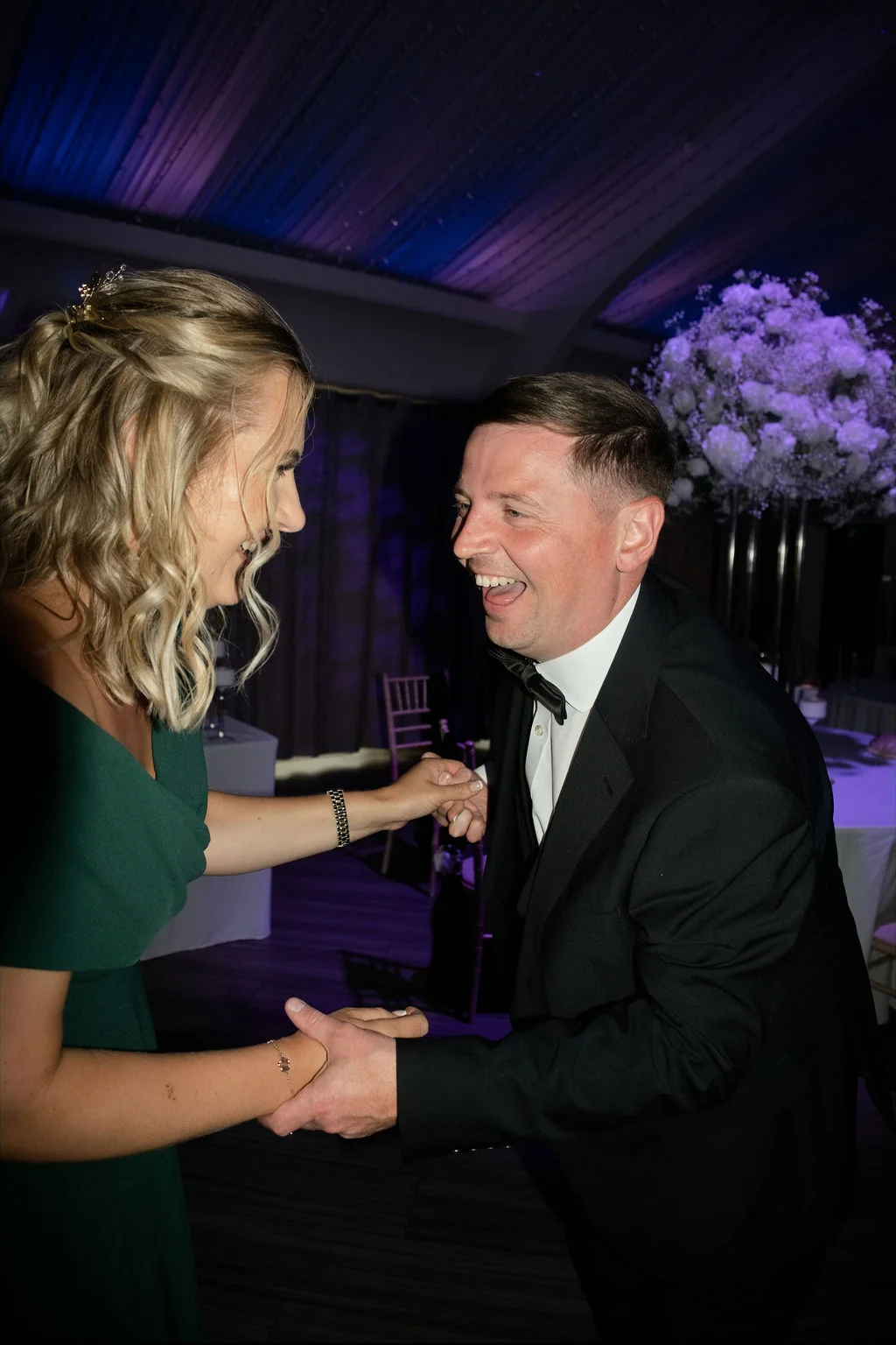 A joyful couple laughs heartily while holding hands on the dance floor during their evening wedding reception.