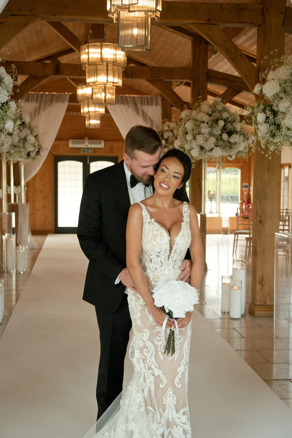A newlywed couple embraces inside an oak-framed barn venue decorated with hanging floral arrangements and warm chandeliers.