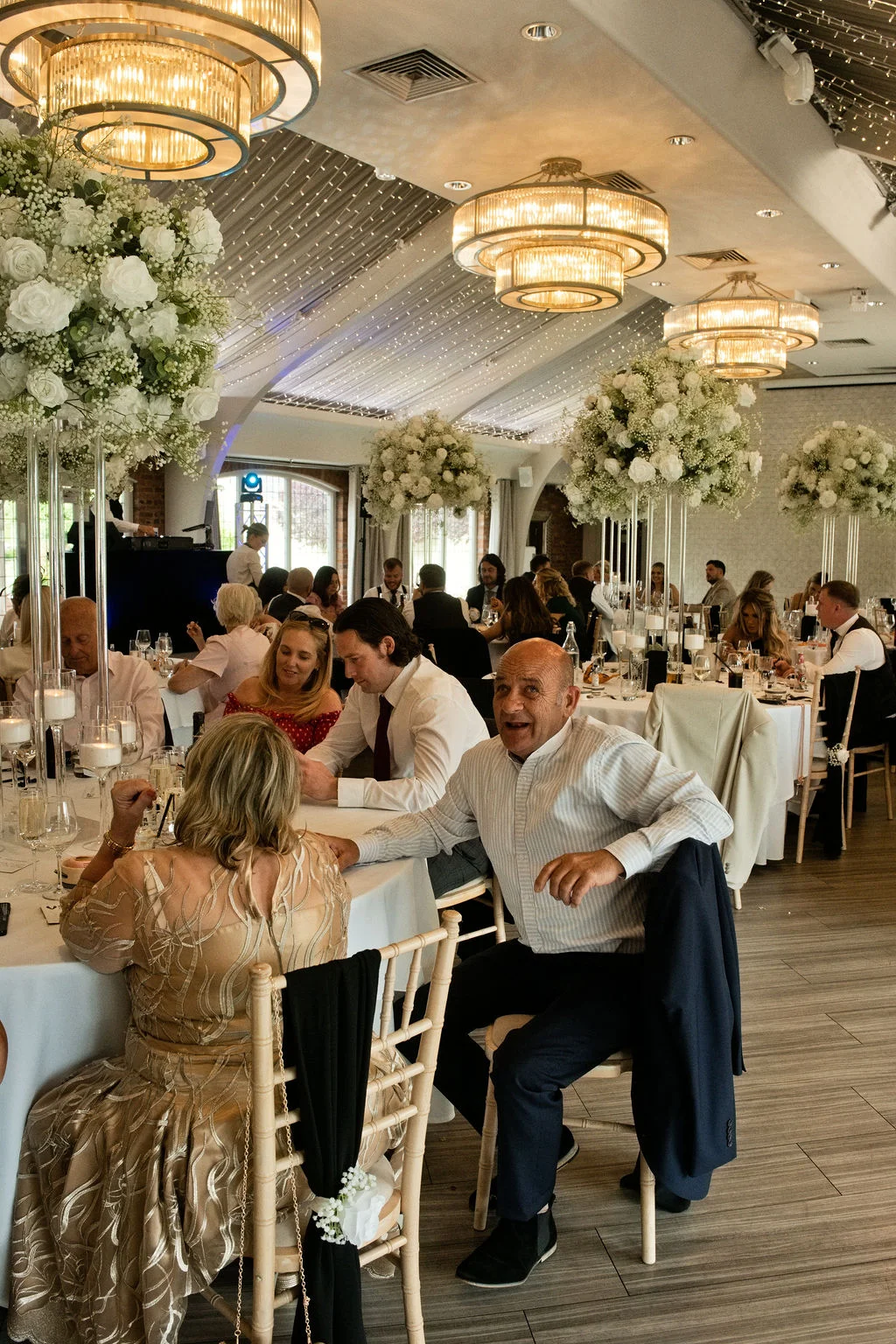 Guests enjoy a lively wedding breakfast seated at elegantly decorated round tables beneath dramatic chandeliers and a sparkling fairy light canopy.