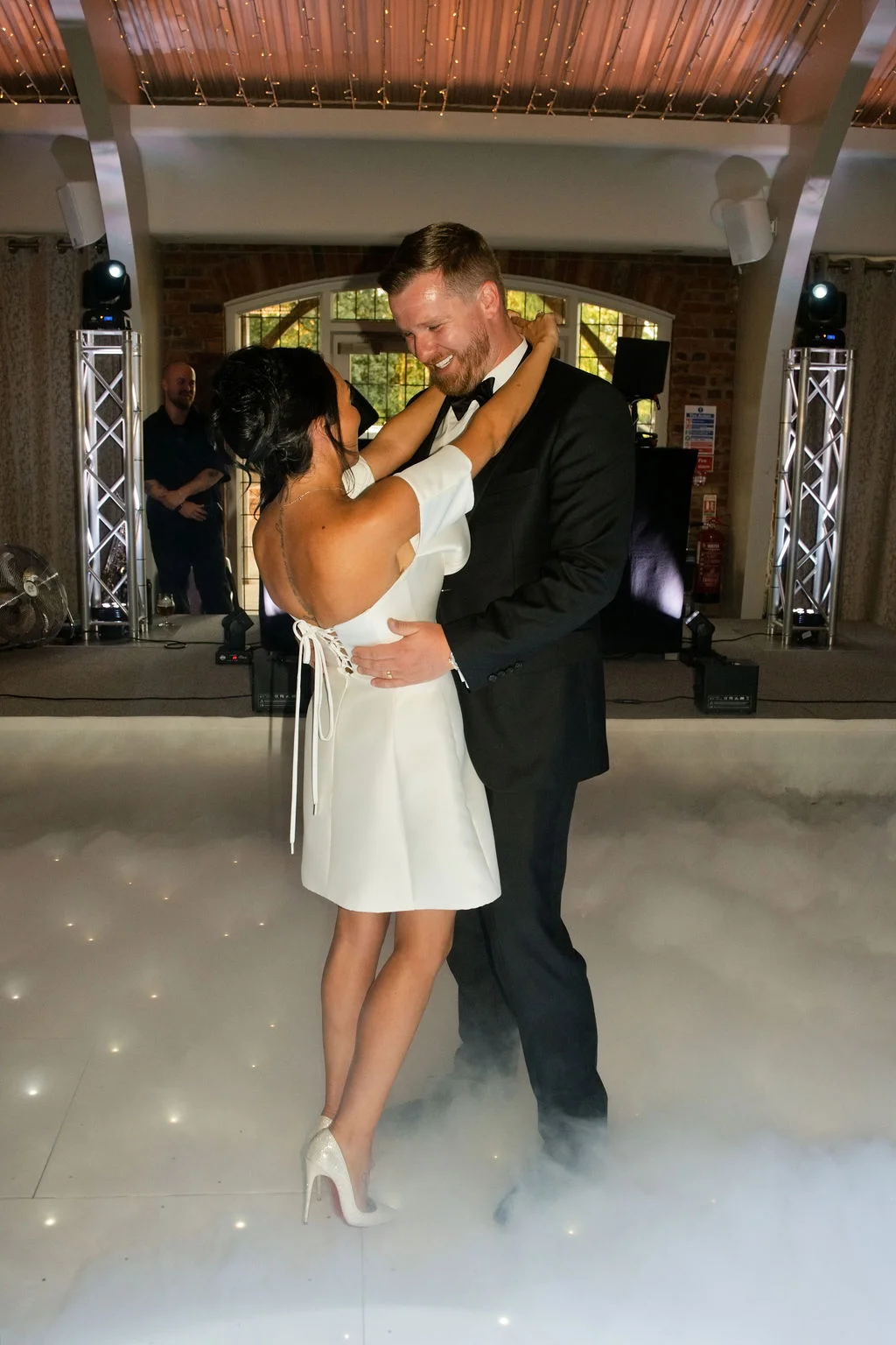 Newlyweds share their first dance on a smoke-filled dance floor, with the bride wearing a short white dress and sparkly heels and the groom in a classic black tuxedo.