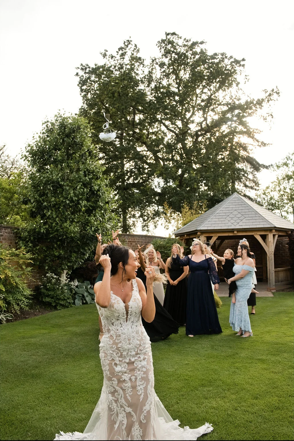 A joyful bride excitedly tosses her bouquet backwards over her shoulder in a lush garden setting while her bridesmaids watch and reach out eagerly.
