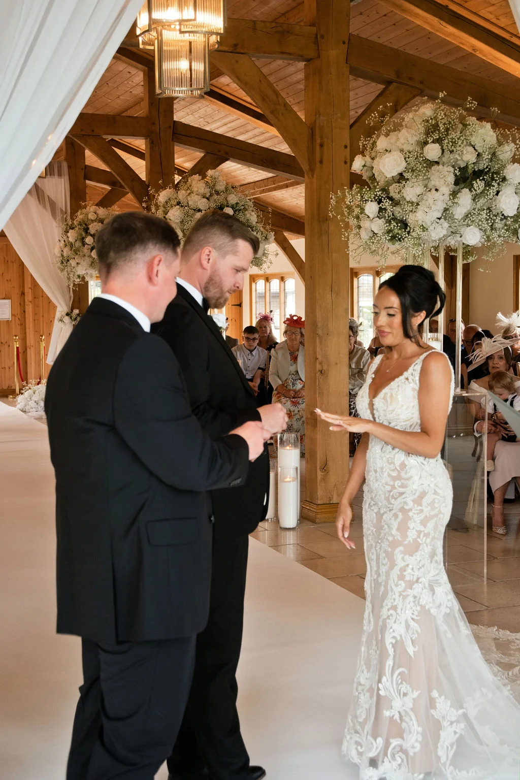 Two grooms in black tuxedos exchange rings with a bride wearing a lace sheath dress during a wedding ceremony inside an oak-framed barn decorated with white floral arrangements.