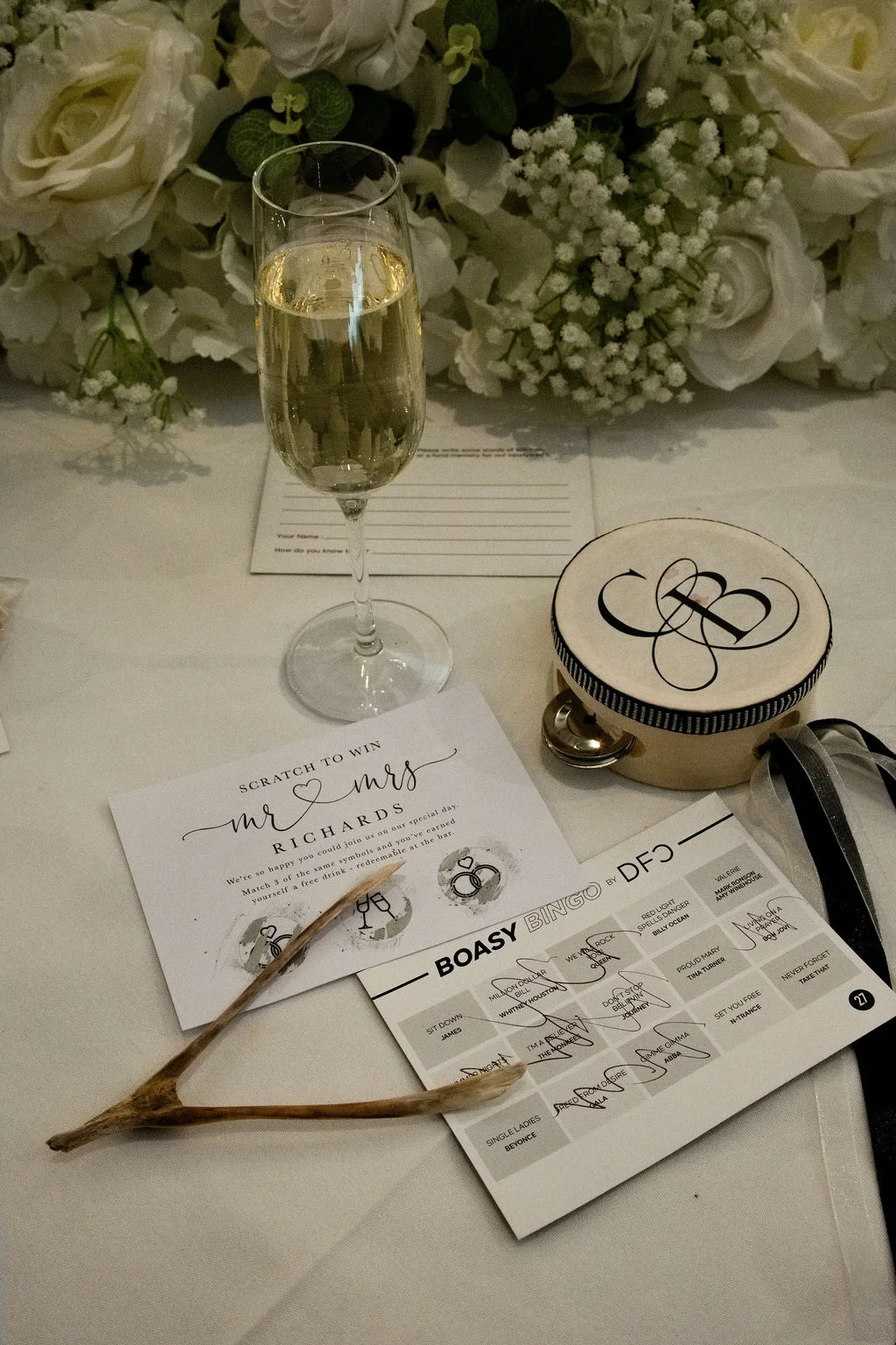 A detailed flat lay shot on a white linen table features a champagne flute, scratch card favours, a custom wedding bingo card, a monogrammed tambourine, and a wishbone beneath a lush floral arrangement of white roses.
