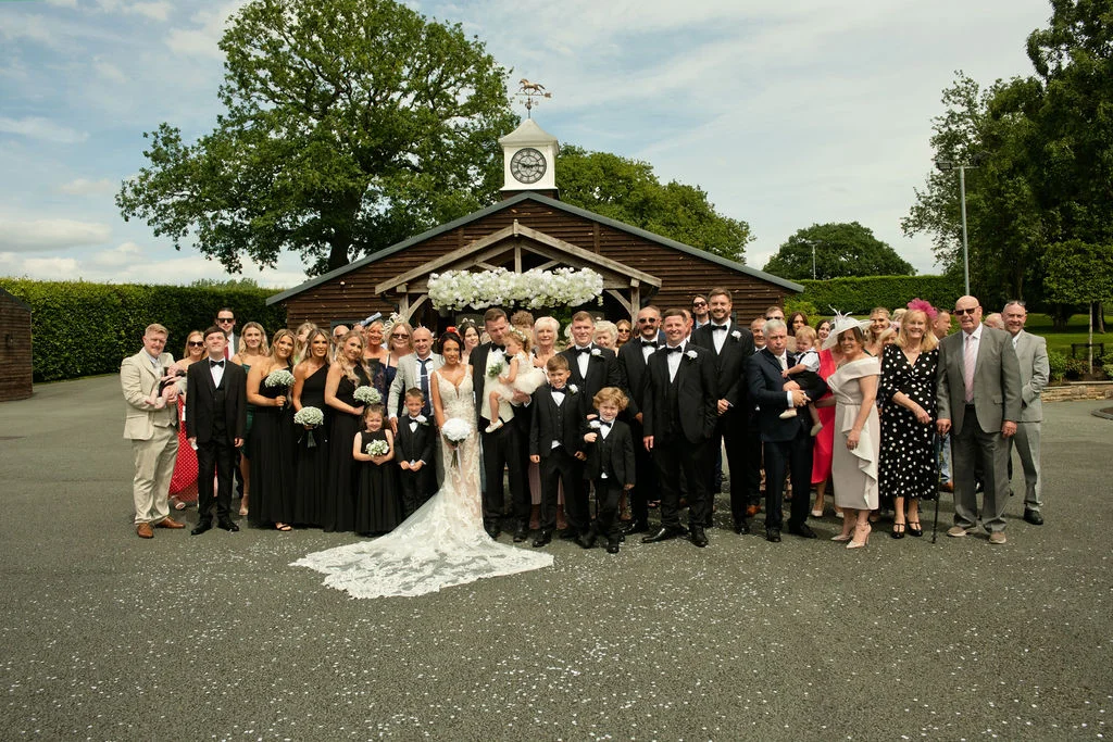 A large wedding party and guests pose for a group portrait outside a rustic wooden barn featuring a clock tower and a floral arch entrance on a sunny summer day.