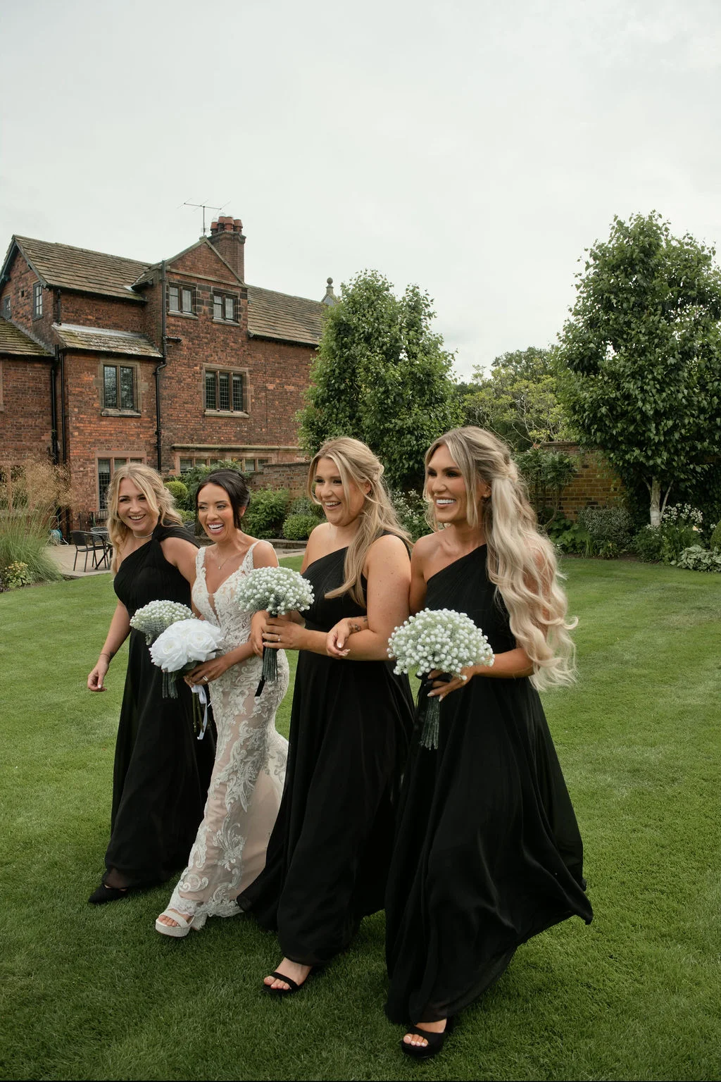 A joyful bride in a lace gown and her bridesmaids in elegant one-shoulder black dresses walk and laugh together across a manicured lawn outside a historic brick country house.