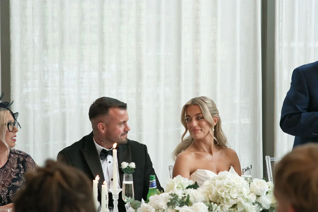 The bride and groom share a tender look at the top table during wedding speeches, surrounded by white floral arrangements and glowing candles.