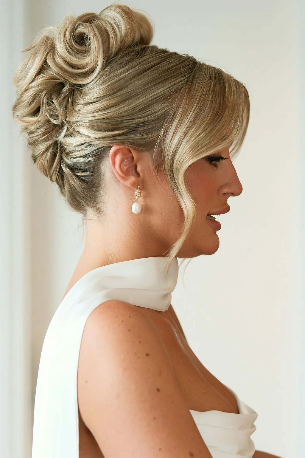 A close-up profile portrait of a blonde bride showcasing an intricate high bun updo, face-framing tendrils, and elegant pearl drop earrings.