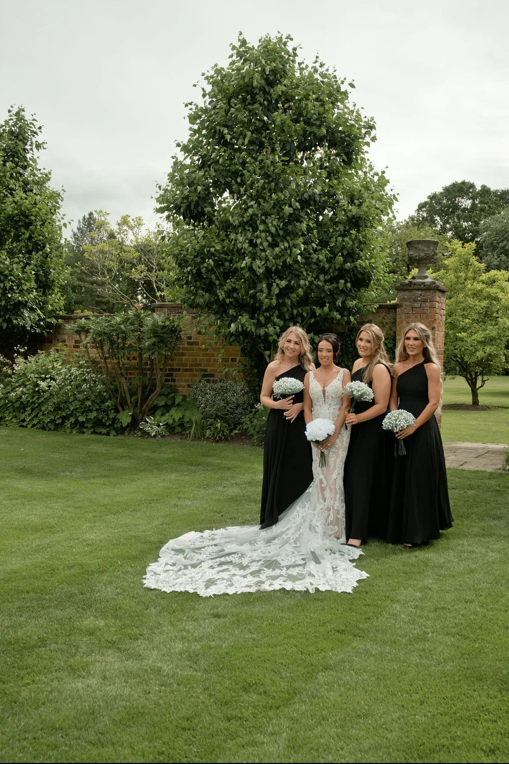 A bride in an intricate lace gown with a long train poses outdoors on a manicured lawn alongside four bridesmaids wearing matching black one-shoulder dresses and holding bouquets.