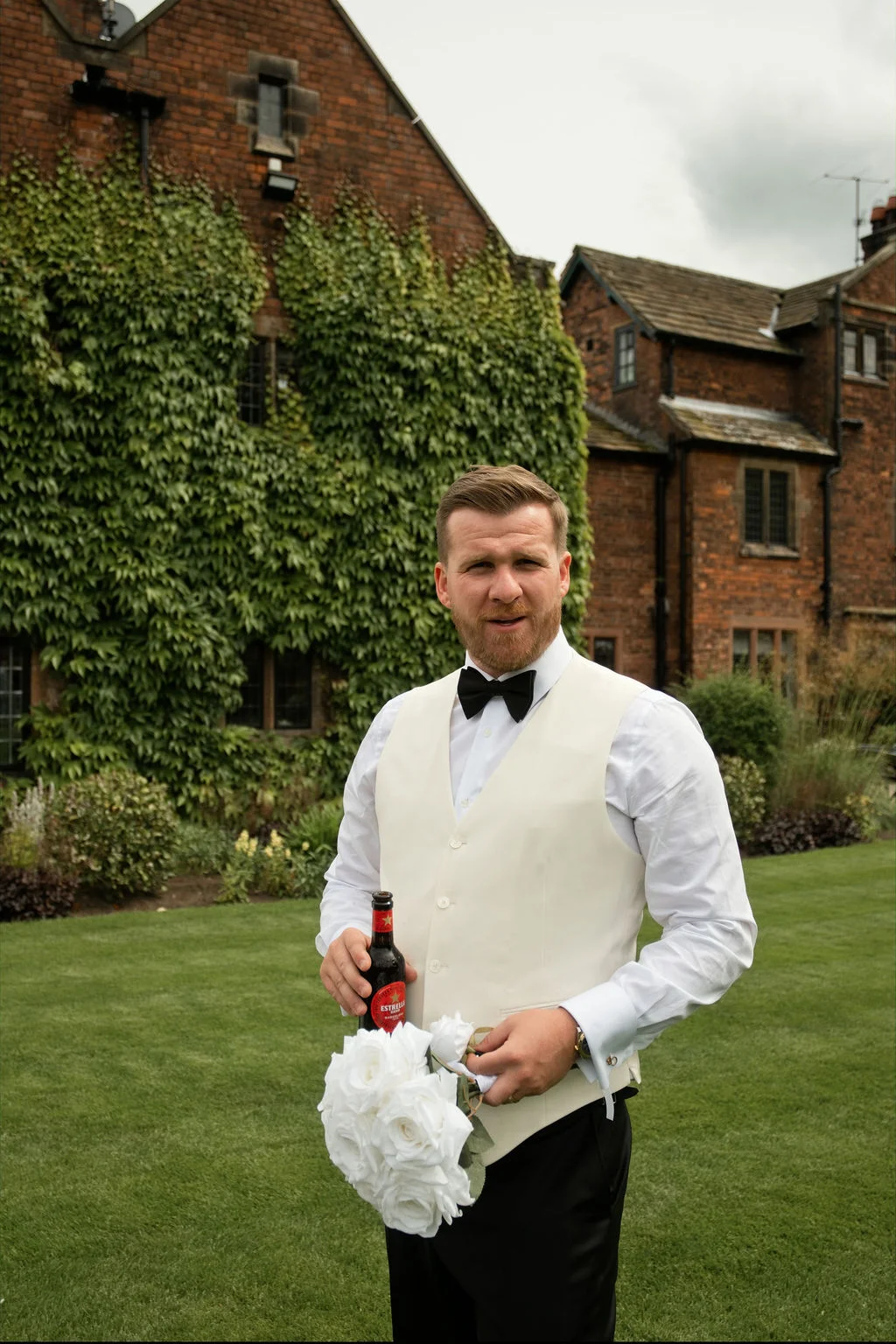 A groom stands on a manicured lawn in front of an ivy-covered red brick manor house, holding a bottle of beer and a bouquet of white roses.