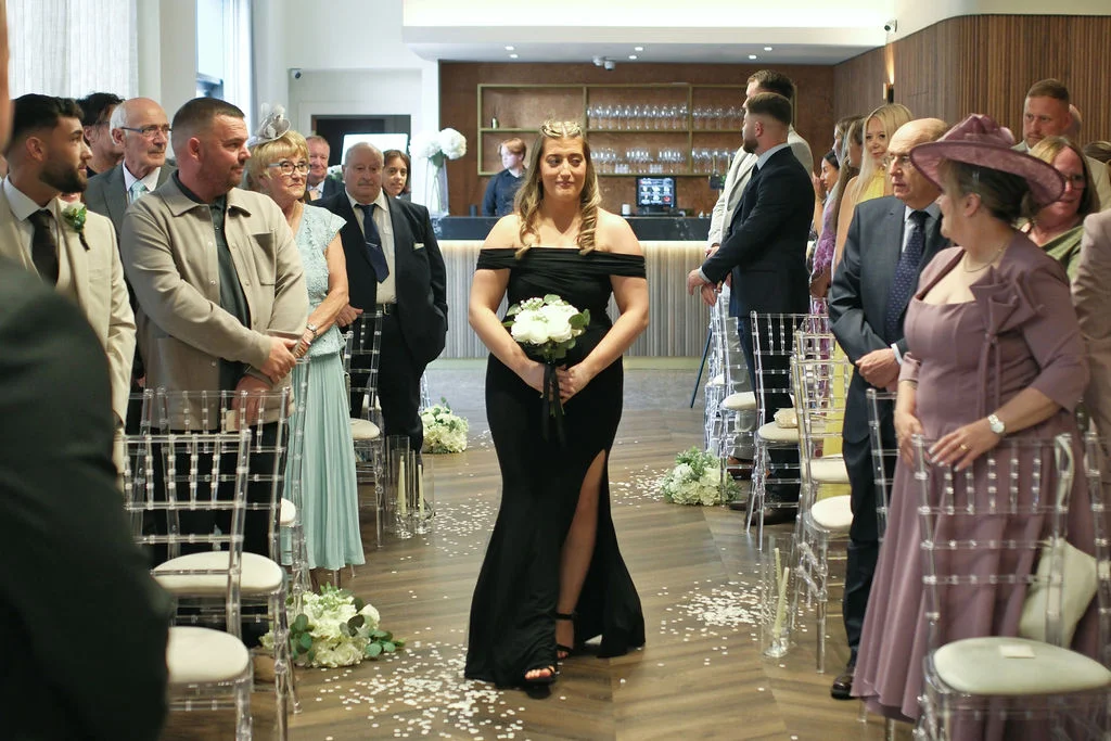 A bride in a striking off-the-shoulder black wedding dress with a high thigh slit walks down the aisle holding a white bouquet while guests watch.