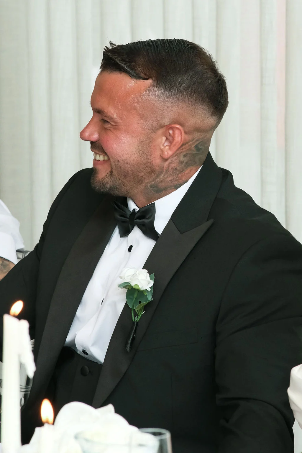 A smiling groom wearing a black tuxedo with a black bow tie and a white rose buttonhole looks off to the side during the wedding reception.