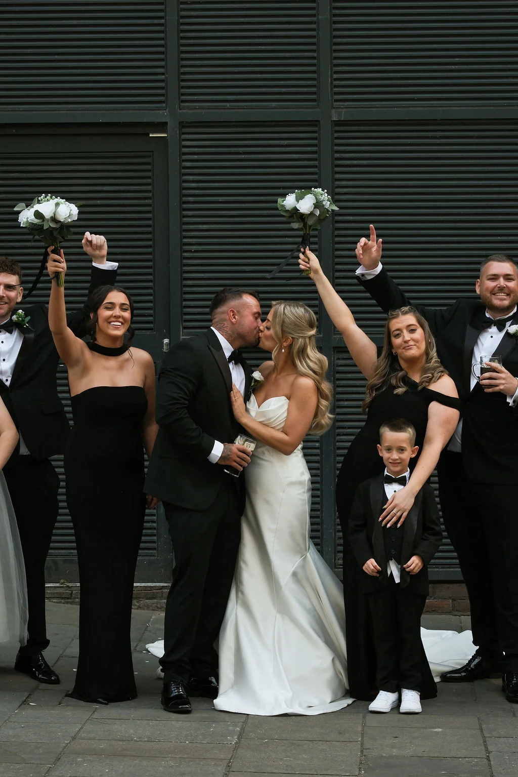 A newlywed couple shares a kiss surrounded by their bridal party dressed in sleek black attire against a dark slated wall backdrop.