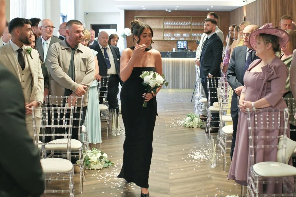 A bridesmaid in a sleek black dress walks down the aisle holding a white bouquet while emotional guests look on.